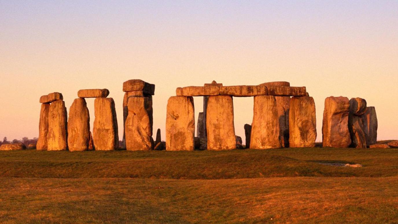 Image of Stonhenge at dusk. Large rectangular stones stand in a circle, some with other stone laid on top to bridge the gap between the stones. The surrounding land is covered in green grass and the horizon is orange.