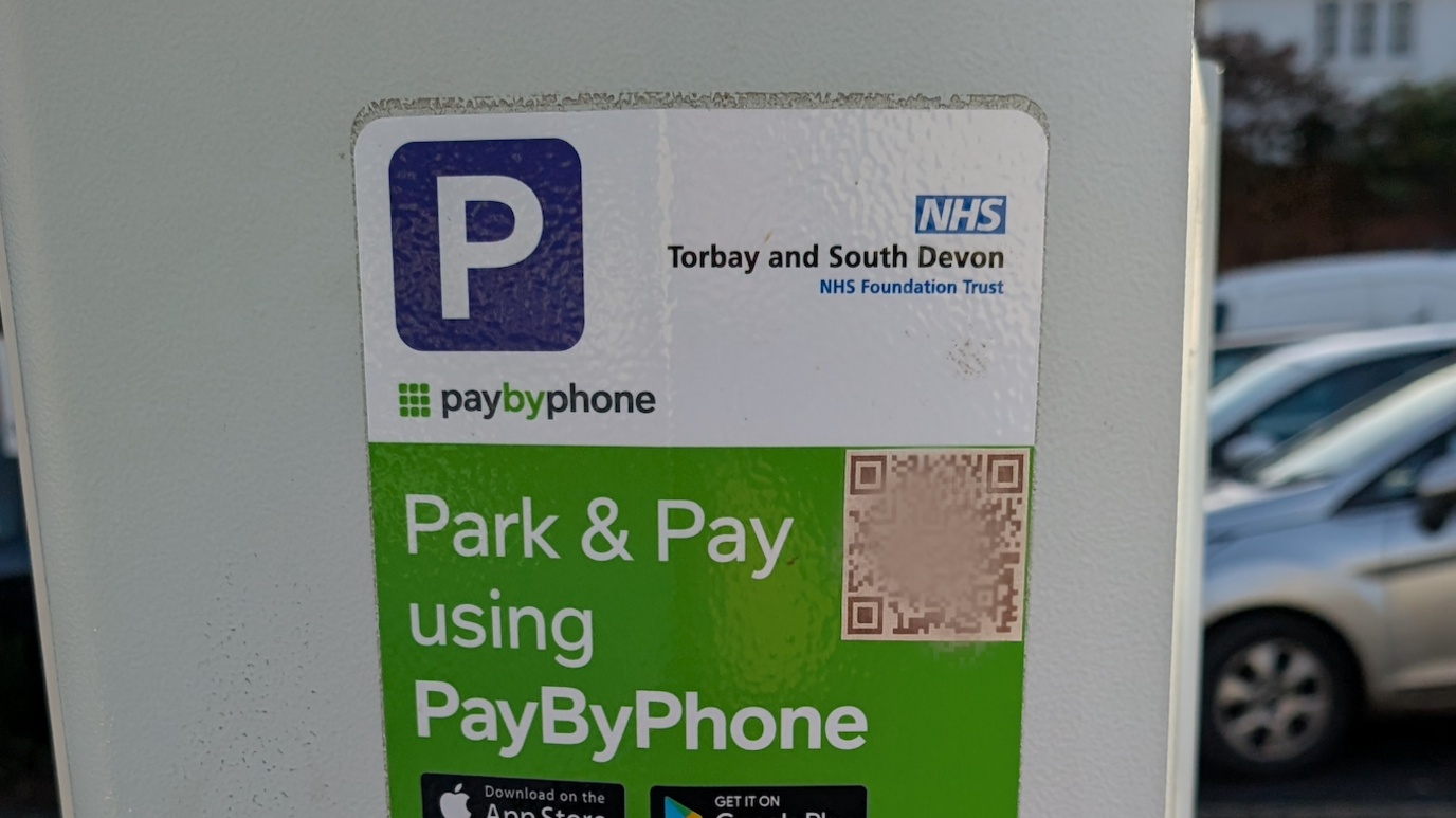 The front of a Park and Pay machine at Totnes hospital with a square QR code image in the top right corner and cars parked behind.