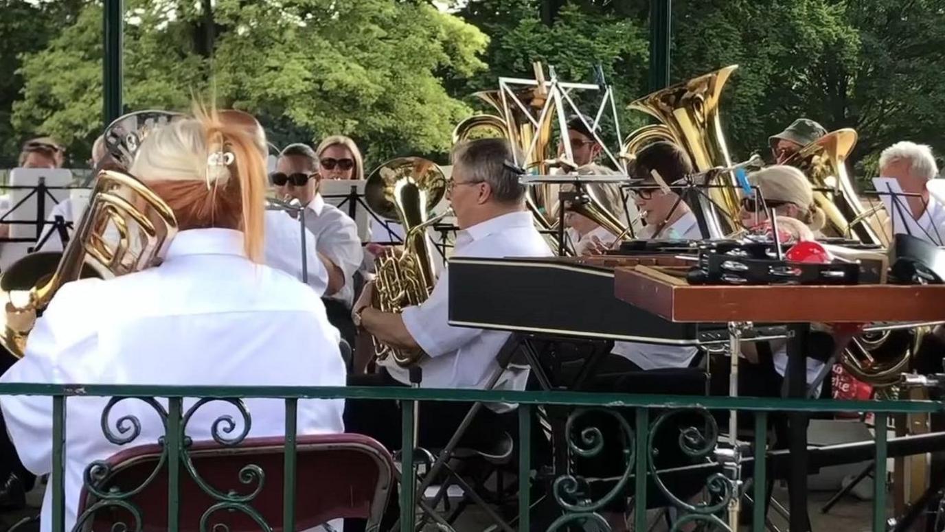 People sitting in a bandstand holding various instruments including trombones, euphoniums, tubas, cornets and trumpets. The musicians are wearing white shirts and dark trousers. A music stand with clothes pegs attached is visible near the camera, along with a xylophone and a glockenspiel.