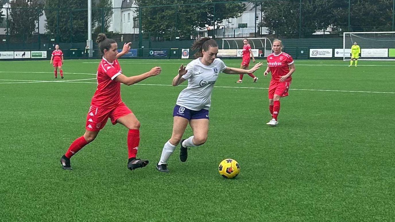A Jersey Bulls player with her hair in a bun and in all-red kit is tracking a Molesey Athletic player in white shirt and blue shorts in the middle of the Molesey Athletic half.
