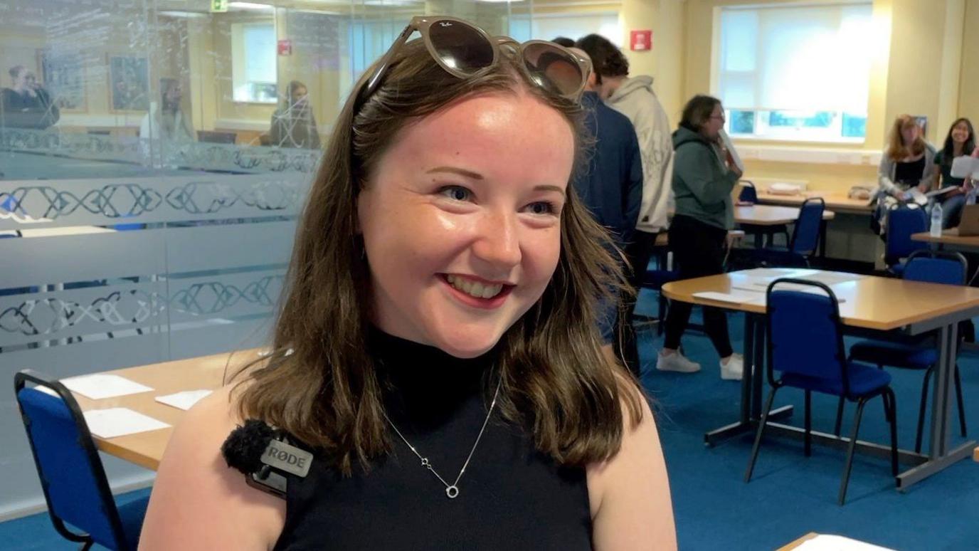 Danielle Wilkinson smiling while wearing a black polo neck and beige sunglasses on her head. There are school chairs and desks and other students in the background.