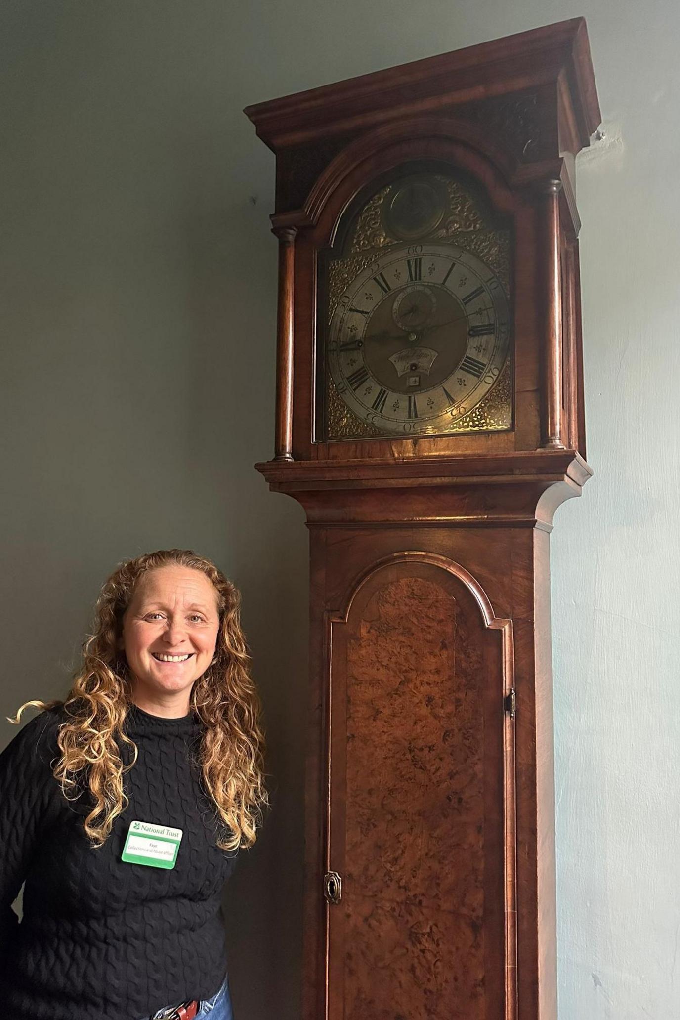 Faye Rason stands smiling next to a grandfather-style clock which towers some 1.5m (4ft) above her. It is made of walnut and has an highly decorated gold-coloured face sitting behind a glass door.