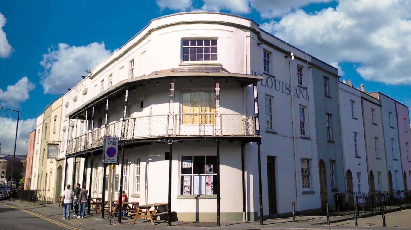 The Louisiana pub in Bristol, taken on a sunny day. It has a 'Mississippi-style' exterior, painted white, with painted iron balconies on the first floor. There are people outside next to benches and tables.