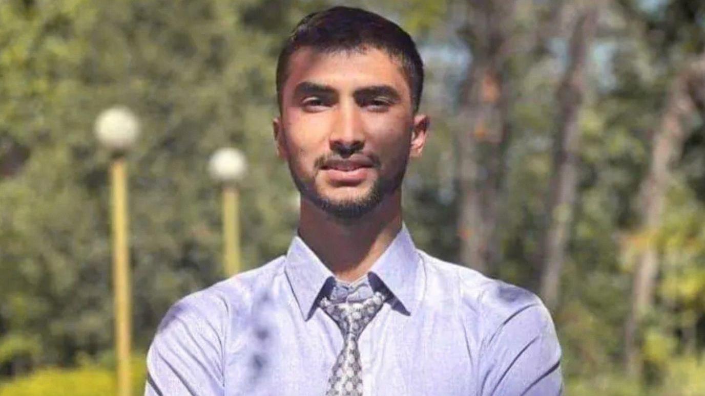 Bipin Joshi, a young man wearing a light blue shirt and patterned grey tie, smiles at the camera in an outside location with trees and lampposts