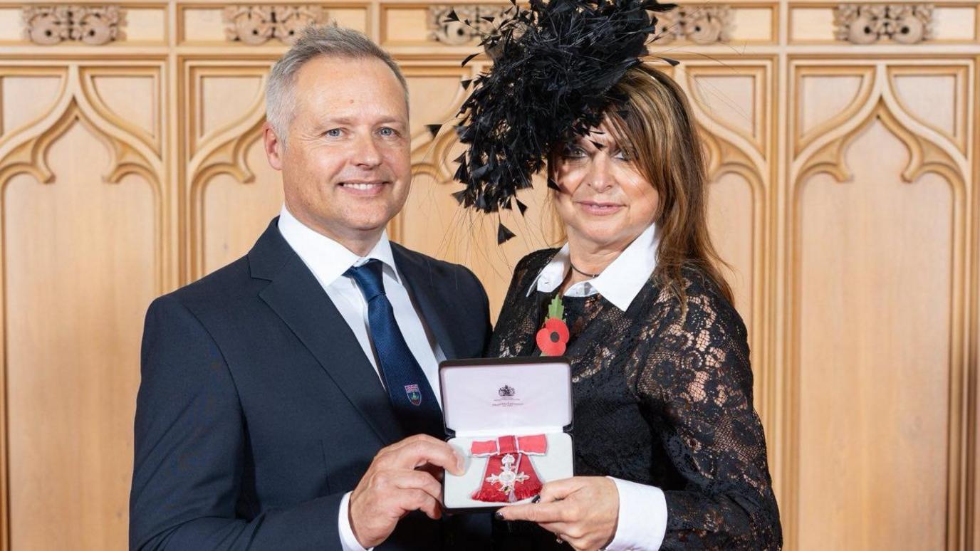 Jo and Andy Priaulx are standing together holding a case with the MBE medal with is a red ribbon bow. She is wearing an elaborate black hat which has a feather effect and a black lace dress which has a white collar and cuffs. He is wearing a navy suit with white shirt and navy tie which has a shield on it. Behind them is wooden panelling.