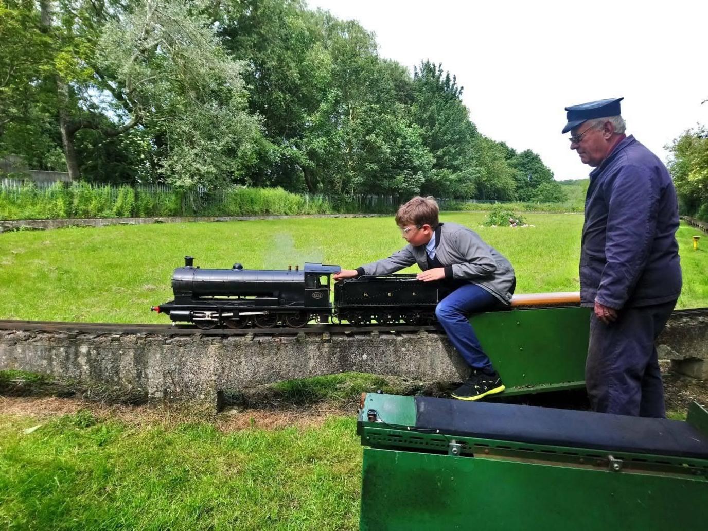 Logan as a boy with a model steam train and carriage. An older man wearing a blue engineer's overall and blue cap watches on. They are outside.