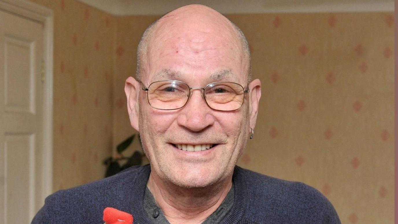 David Newton, wearing a grey sweater over a button-up T-shirt, and metal-framed spectacles, smiles as he holds up a red presentation box embossed with a gold National Blood Service logo and the figure "100" in one hand. In his left ear is an earring. In the other hand is a red soft toy mascot of a blood drop. He is inside what looks like a living room.