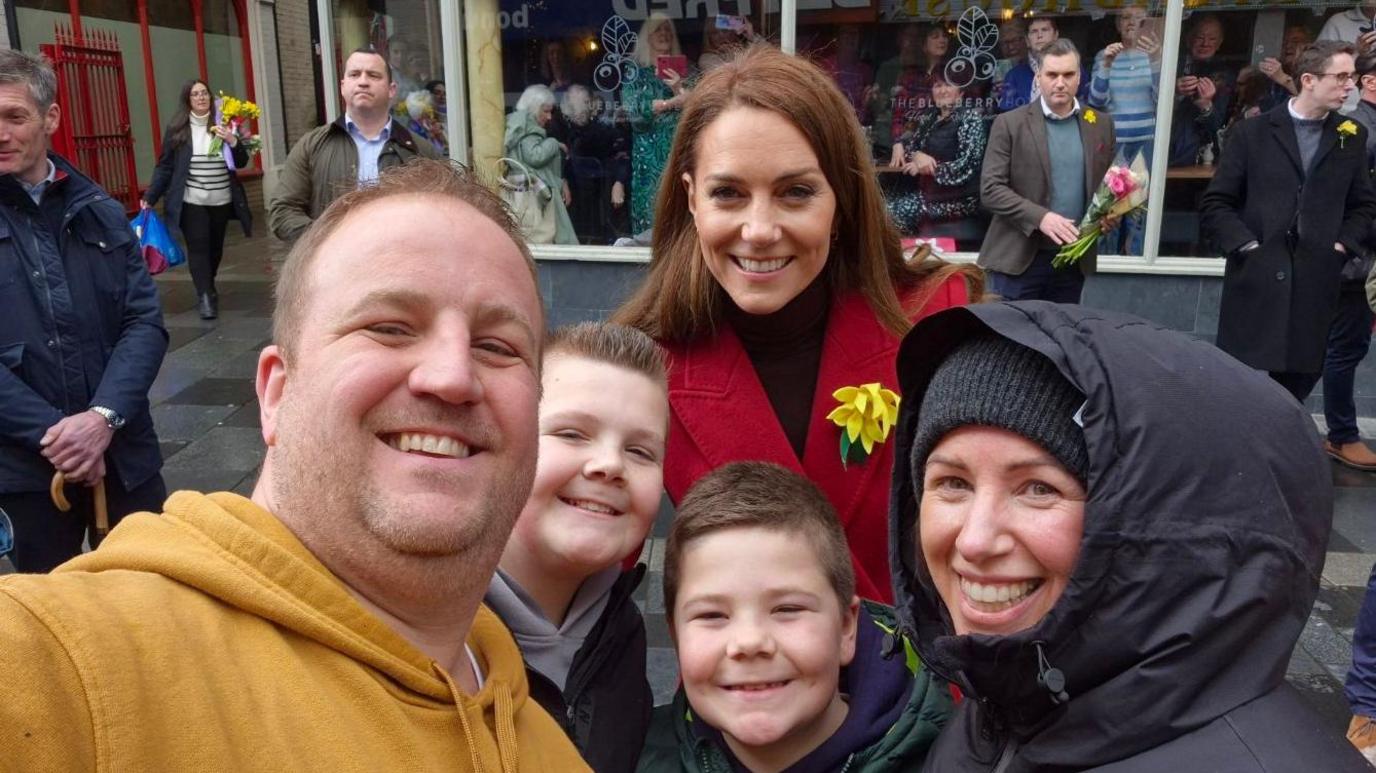 Selfie of Gavin Hailes, 41 who is wearing a mustard yellow hoodie, his son Ralphie, 12, who is wearing a black jumper and has short spiked hair, Joseph, 8, who is wearing a green Puffa jacket, and aunt Hannah Jones, 45. Princess Catherine smiles behind them. 