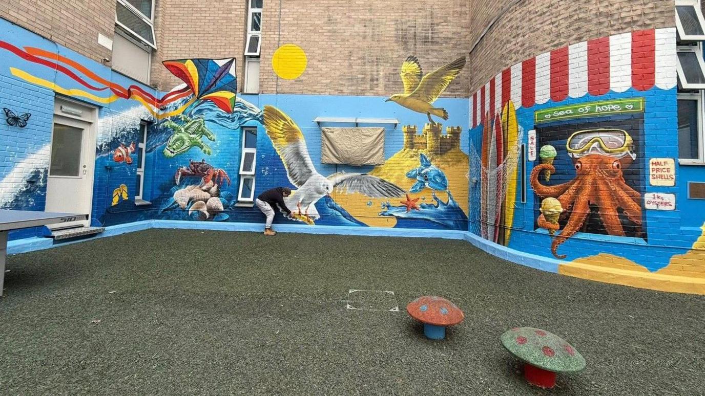 A mural in a hospital garden, which includes a blue background depicting the sea, sea life, sand castles and kites.