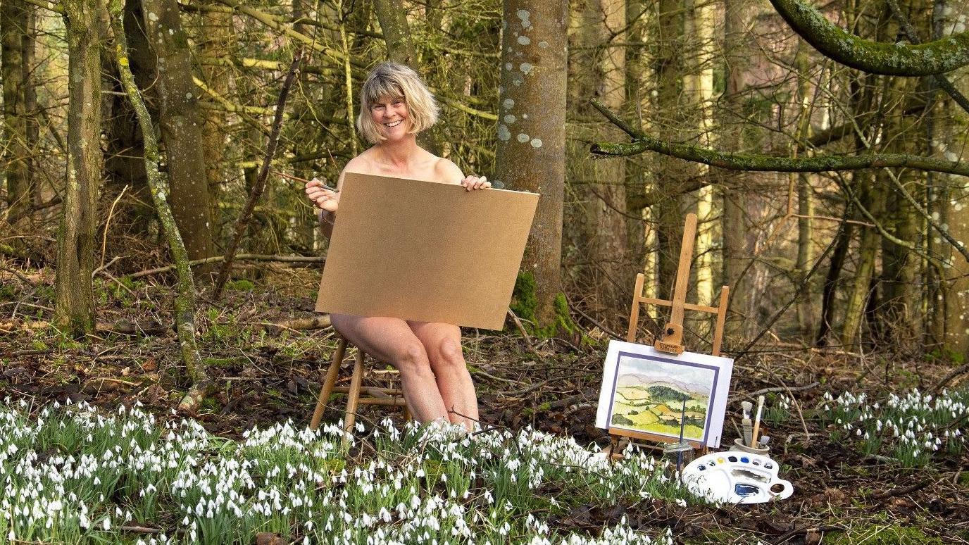 A woman of about 60 with a blonde bob sits on a stool. She is naked but appears to be painting and the board covers much of her body. Next to her are paints and an easel.