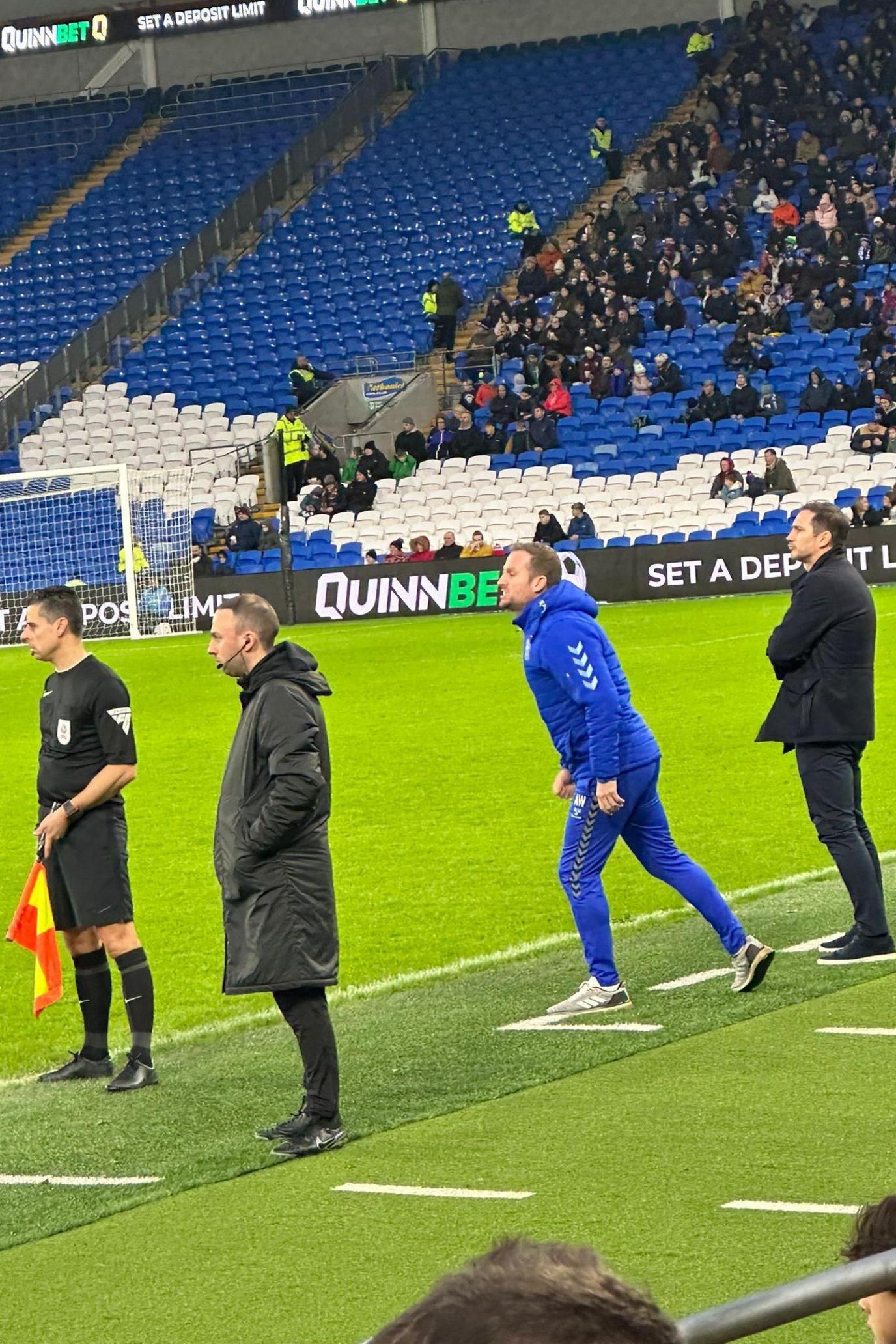 Dale Randall on the sidelines at a Cardiff City vs Coventry game. He's wearing a black coat, black trousers and black trainers. 
He is also wearing an earpiece and microphone