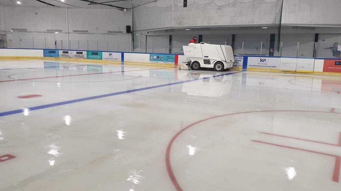 An ice rink is empty with barriers around the side with adverts on. You can see a person driving a white ice resurface vehicle around the edge of the rink.