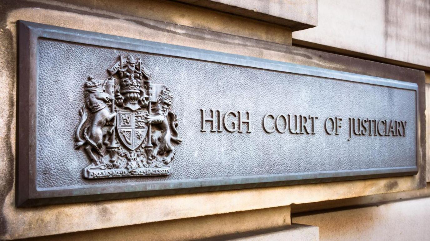 A sign on a stone wall of the High Court in Edinburgh. It has a royal crest and the words: "High Court of Justiciary".