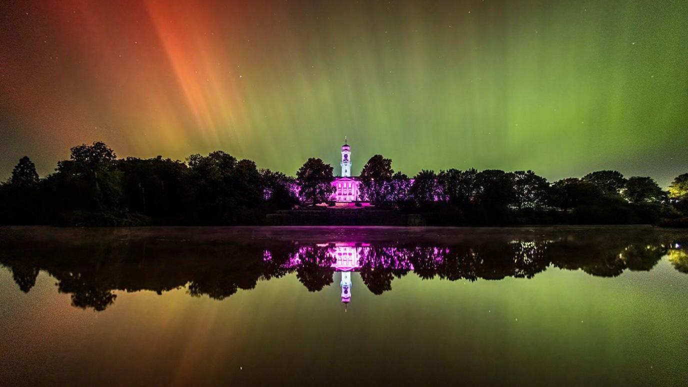 The Northern Lights are shining in green and red over the Trent Building at the University of Nottingham. The picture shows the reflection of the lights in a lake in front of the building.