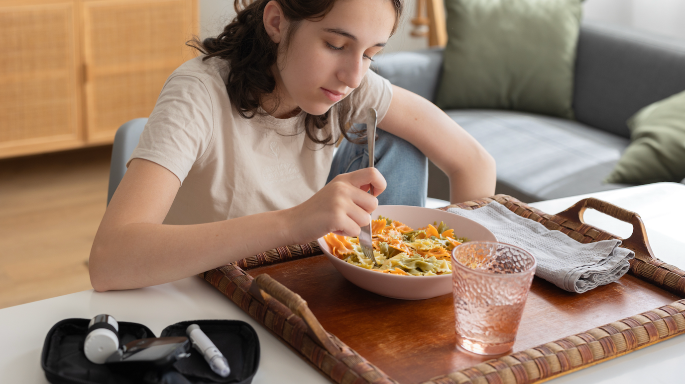 Teenager girl with diabetes is eating pasta at home and checking her glucose level with a glucometer