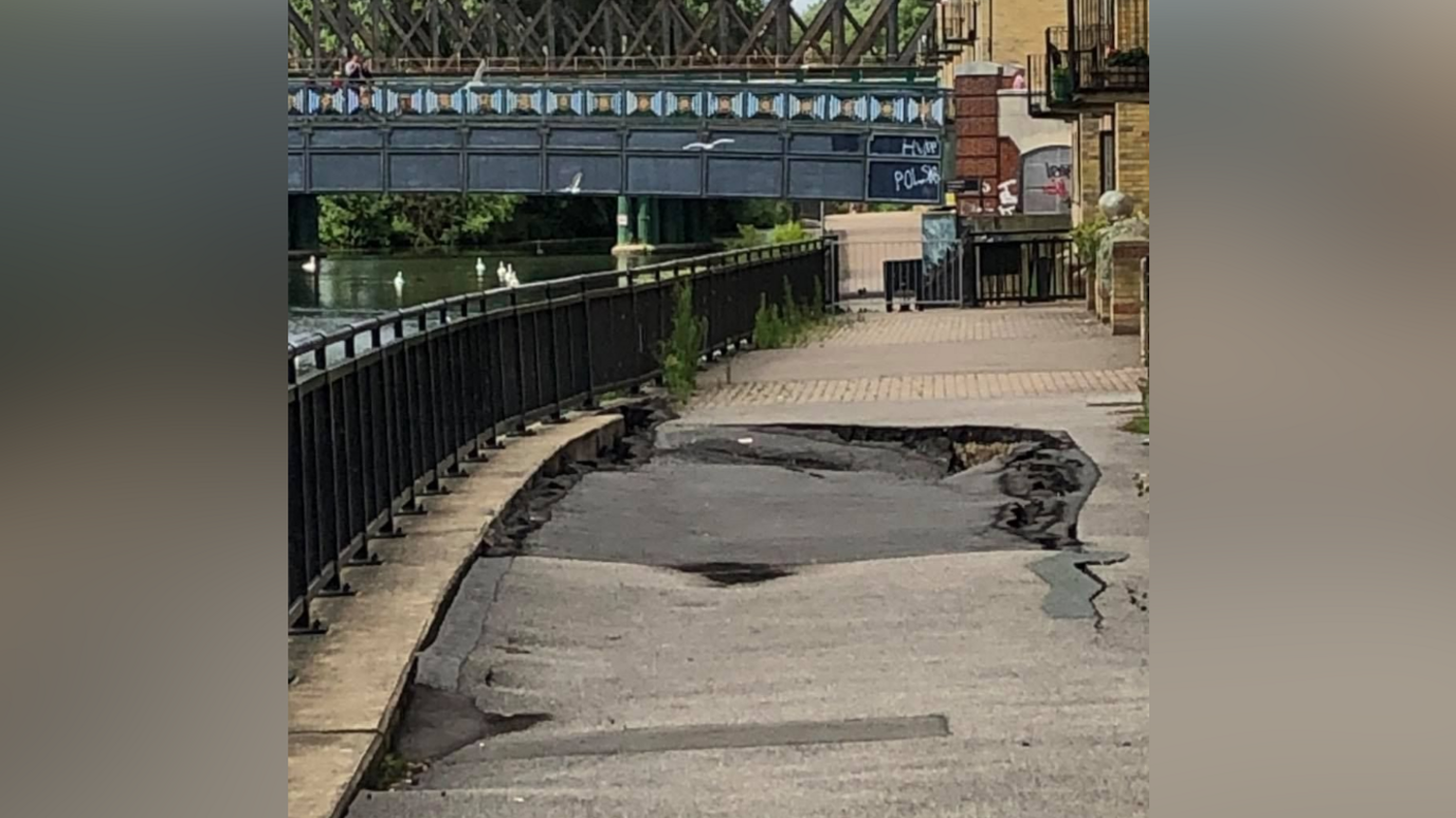 A tarmac pathway running beside a river which seems to be buckling and  has a small hole in the middle of it. A blue iron railway bridge can be seen in the background