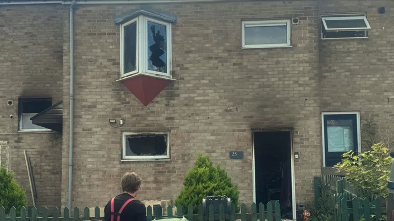 The outside of a house. The windows are blown out and the front door is open with signs of smoke damage.