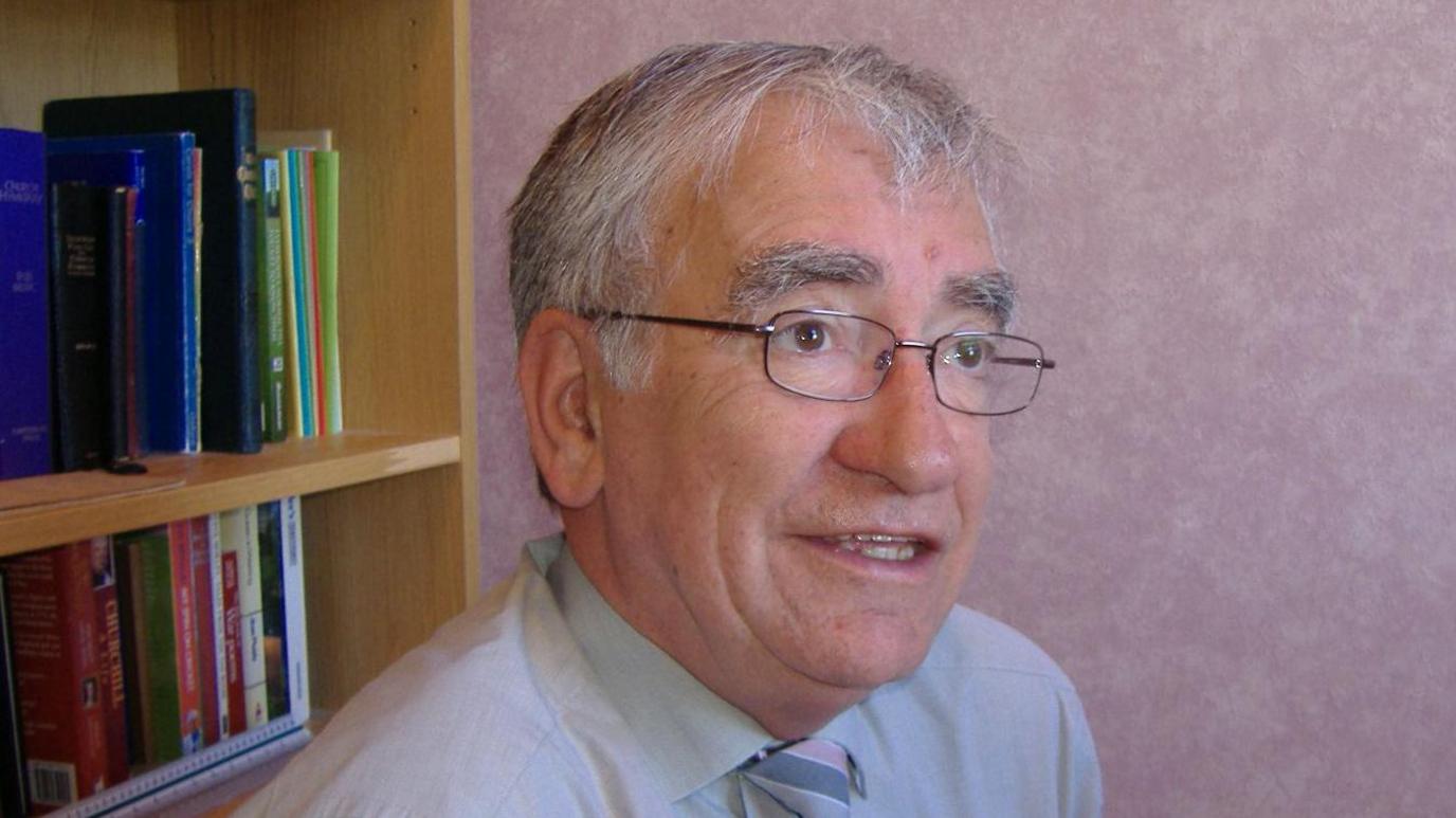 A grey-haired man with glasses, wearing a grey shirt and striped tie, looks beyond the camera. In the background is a bookcase