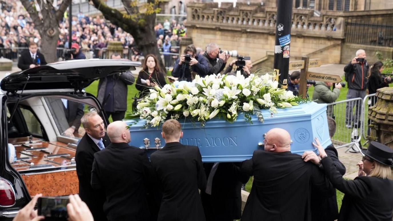 A blue coffin, which is adorned with white flowers, is carried out of Manchester Cathedral by Ricky Hatton's family and put into the back of a hearse. Crowds of people can be seen paying their respects in the background.