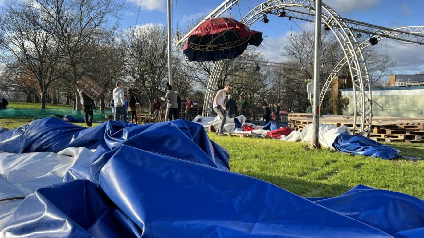 Blue canvas lies on the ground with tent construction metalwork and a hood in the background with a number of people working.