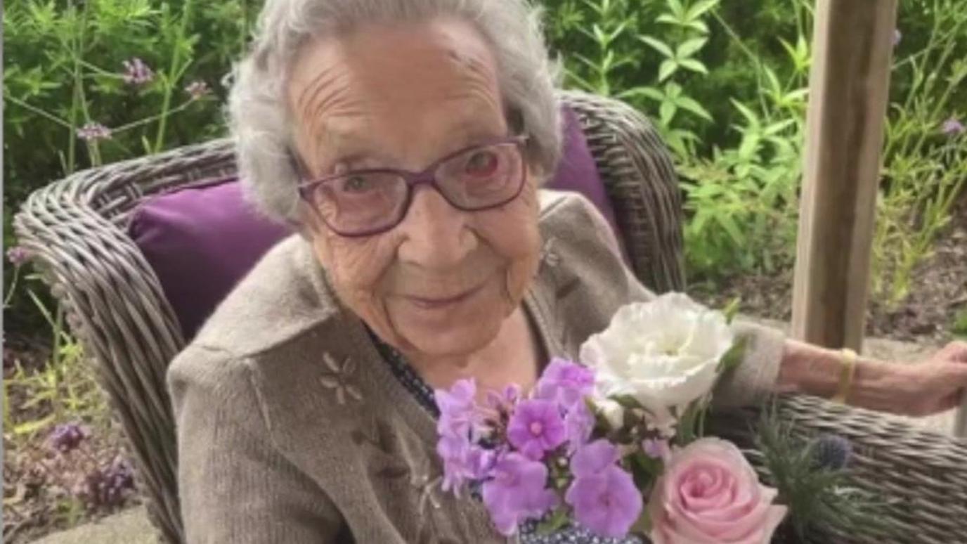 Betty, a woman with short grey hair and purple glasses, sitting in a wicker chair with a purple cushion. She is holding a bunch of pink and white flowers in one hand while grabbing a walking frame out of shot with her other hand. There is greenery in the background. 