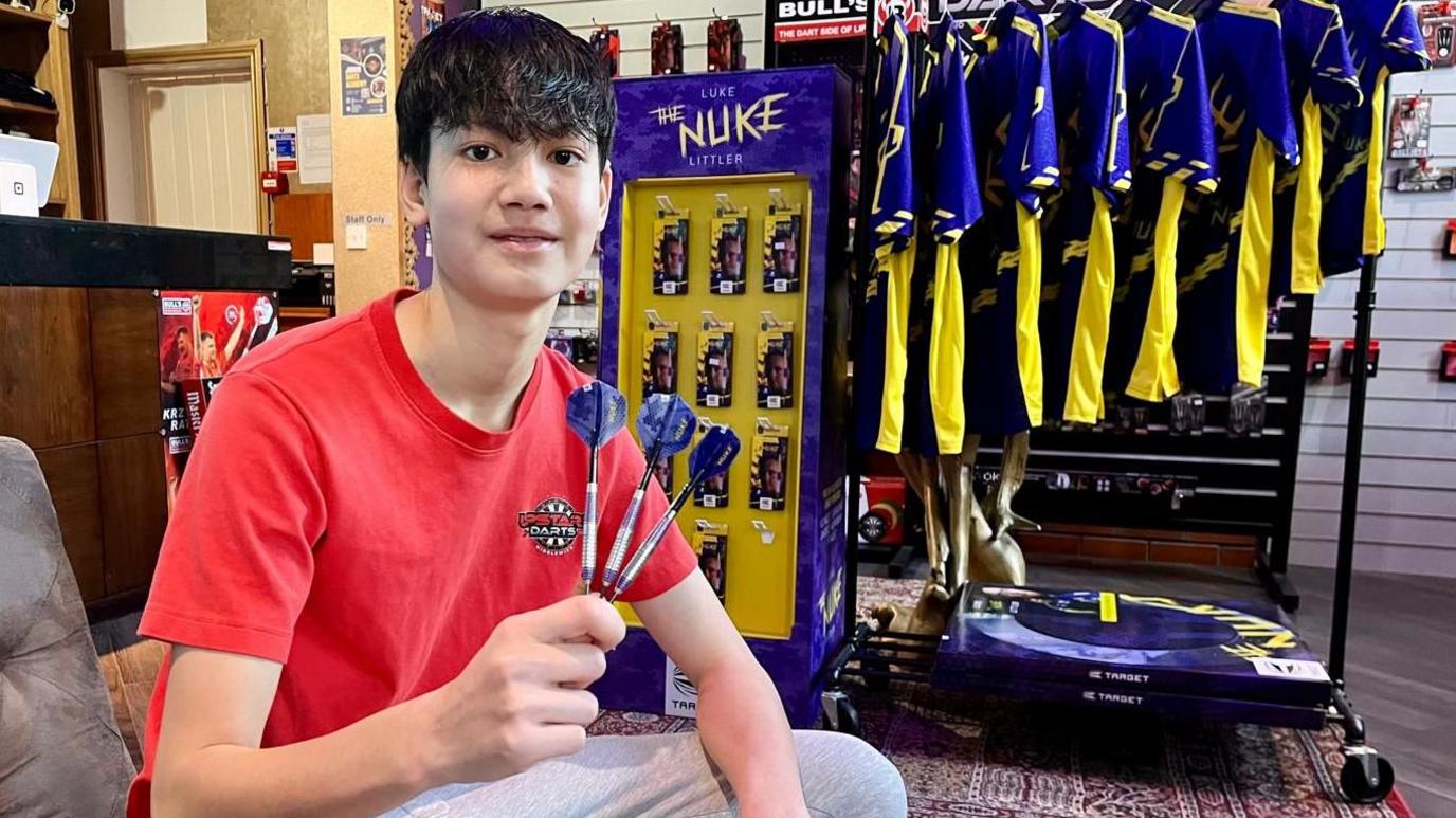 A teenage boy sits on a brown sofa in a darts equipment shop. He is holding three darts in his hand. The darts are dark blue and with the name 'Luke The Nuke Litter' written in yellow, as are the shirts, display unit and darts boards displayed behind the young man. He is wearing grey tracksuit bottoms and a red t-shirt with a darts logo on the left chest.