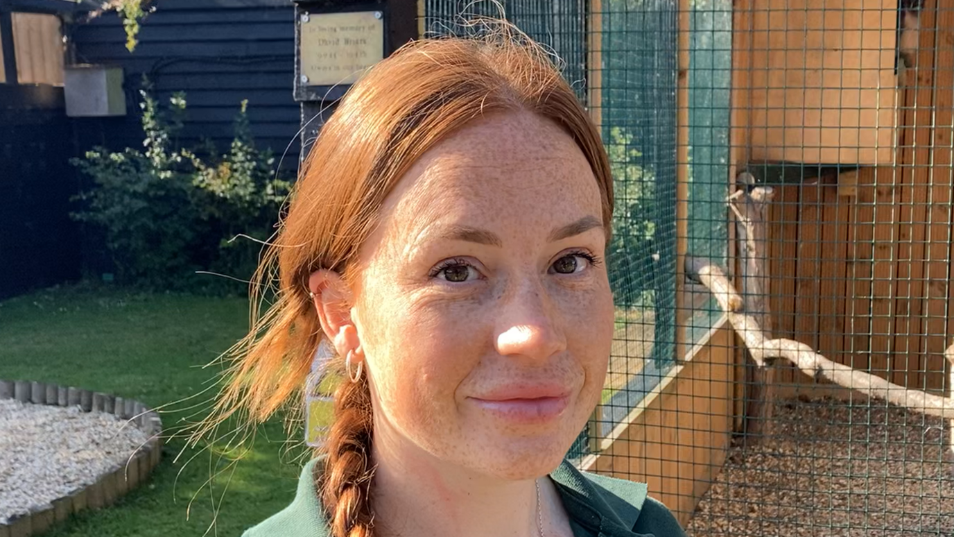 Emily Quantrill smiles at the camera in front of an enclosure. She has red hair that has been tied back and braided, with the braid resting over one of her shoulders.