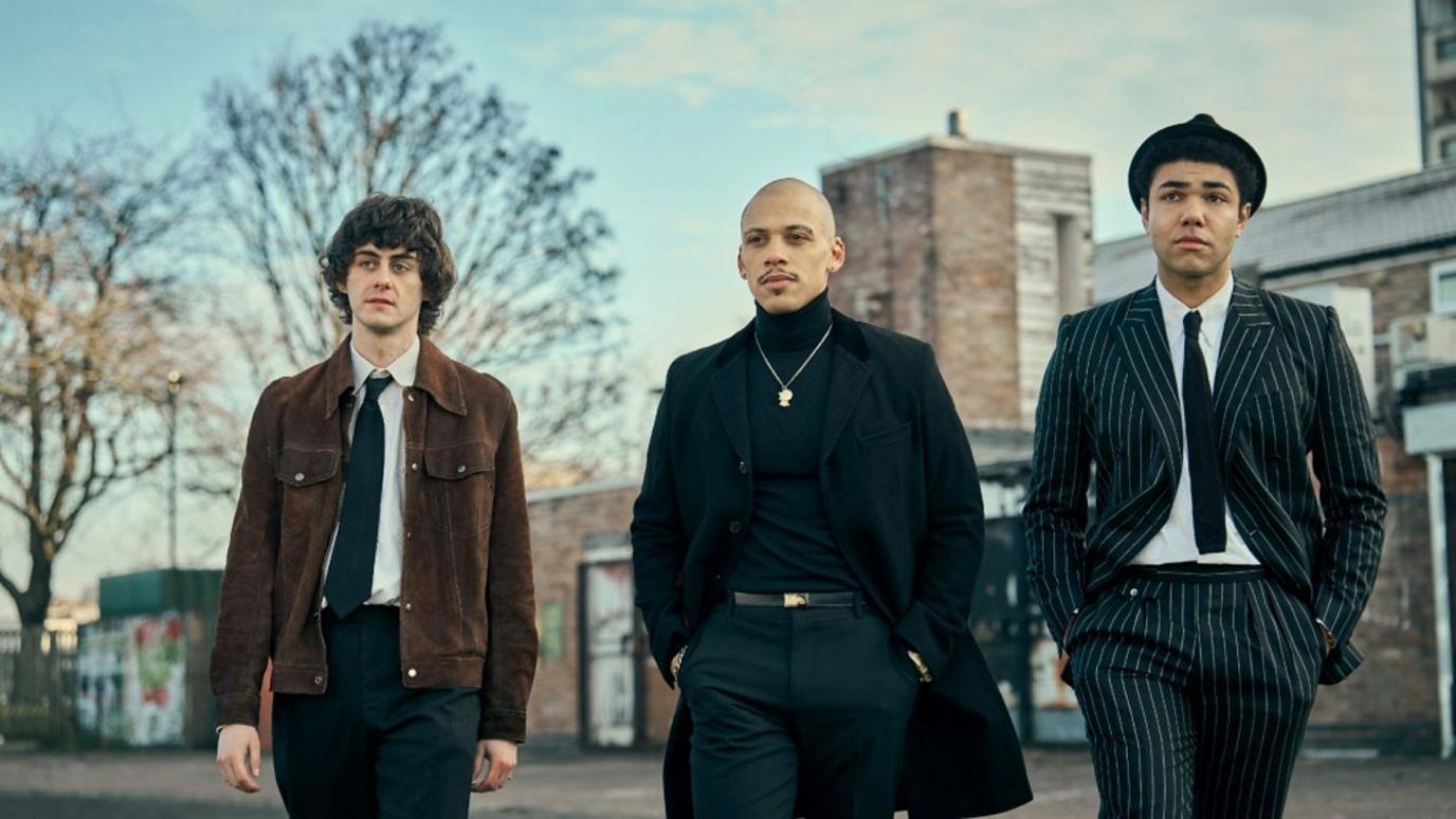 Three men crossing a street. The man on the left is in a brown jacket, black tie, white shirt and black trousers. The man in the centre wears black trousers, polo neck and long coat and has a silver pendant necklace. The man on the right is in a pinstripe black and white suit with white shirt, black tie and black hat.