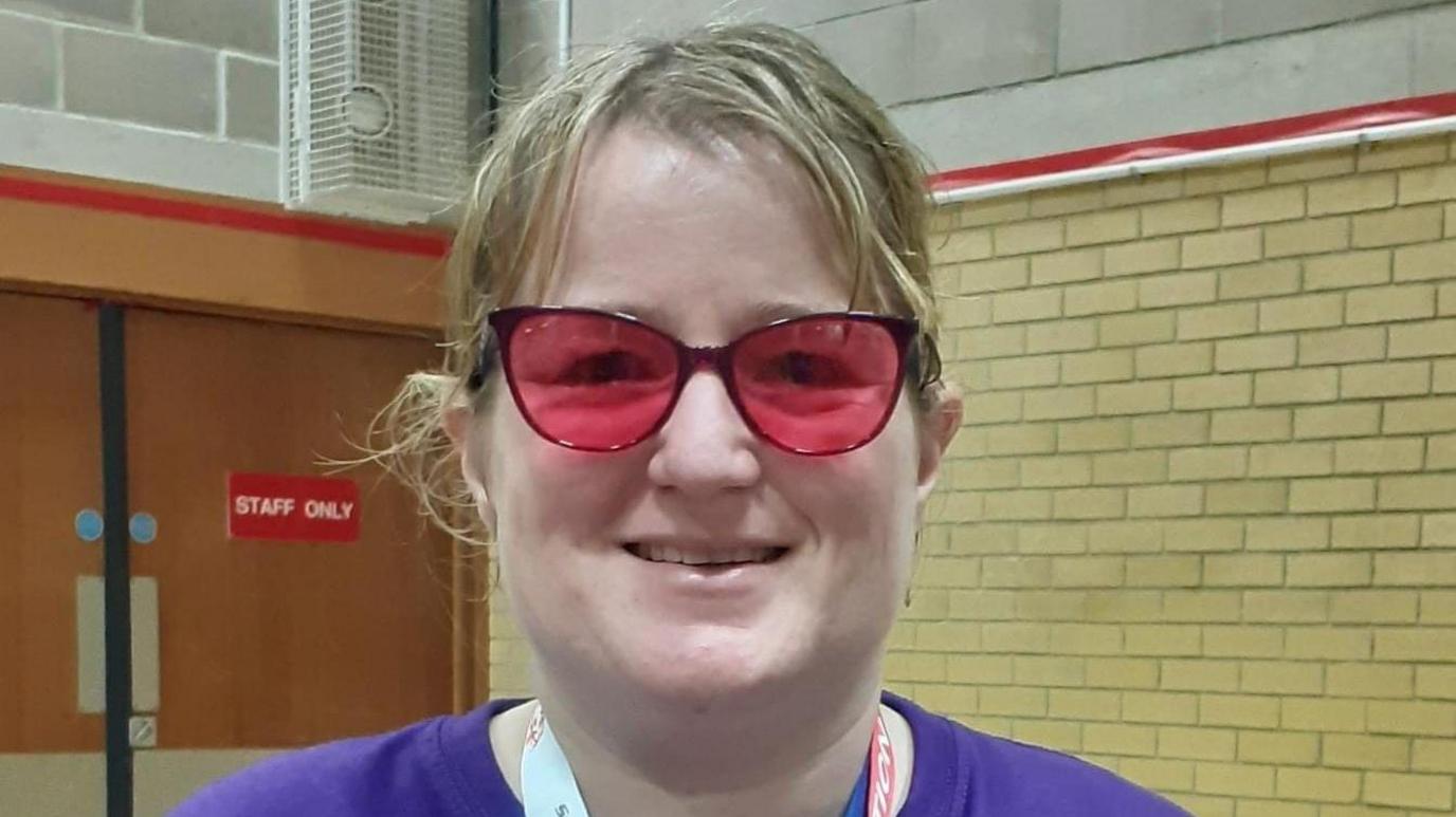 Donna Wyatt smiles at the camera while standing in a sports hall. She wears pink tinted glasses and a purple t-shirt.