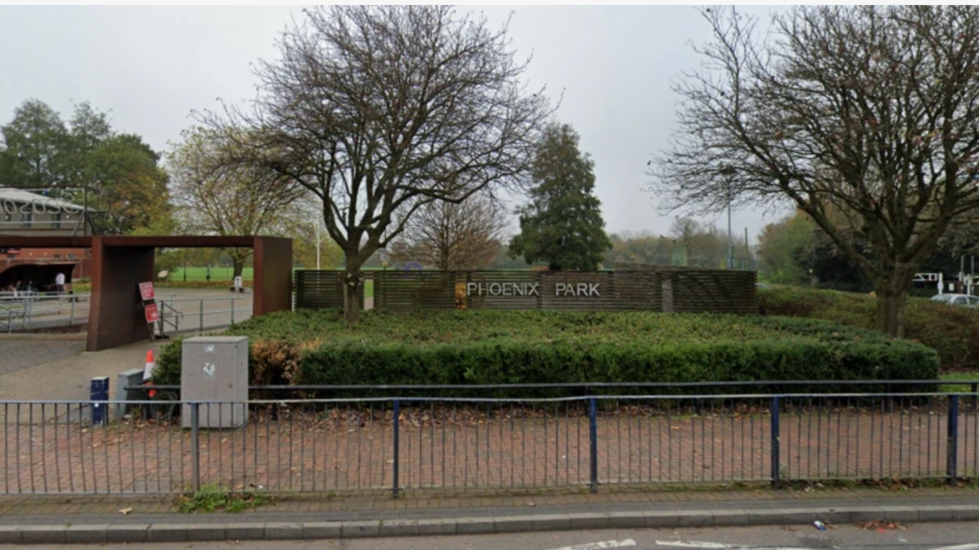 The park entrance is on a main road and can be seen behind some road-side railings. The name of the park appears on a wooden fence with the entrance either side of it and low hedge in front of it.