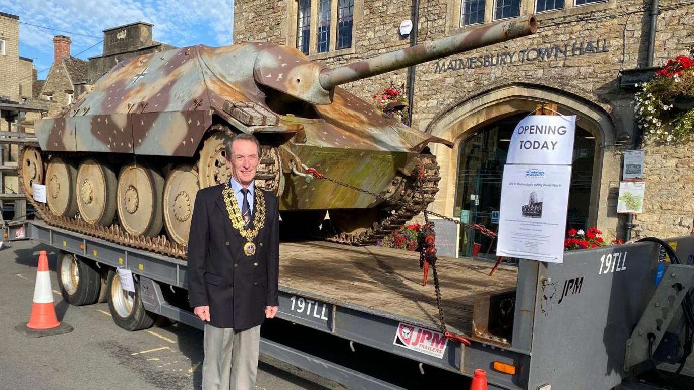 A mayor in a gold chain wearing a suit smiles at the camera while standing in front of a historic tank which is on the back of large trailer. Malmesbury Town Hall, a Cotswold stone building, is visible in the background.