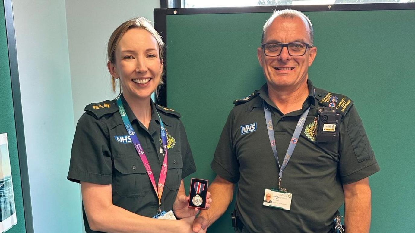 Claire Ruddy shaking Stuart Little's hand as she hands him a small medal. Claire has a multi-coloured lanyard around her neck, blonde hair and is smiling. Stuart has a blue lanyard around his neck, is wearing glasses and smiling. Both are wearing green ambulance uniform.