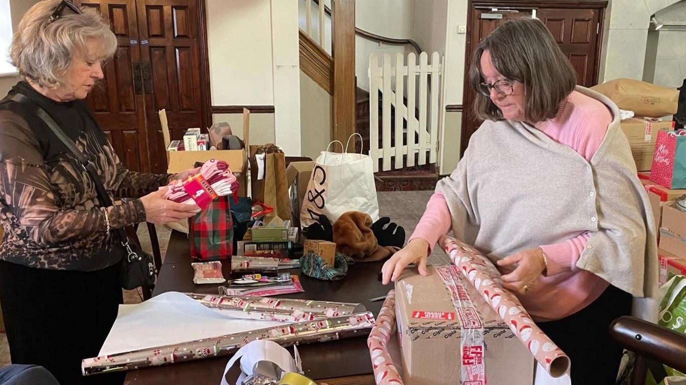 Two women pack presents on a table in a hall for the Ukraine