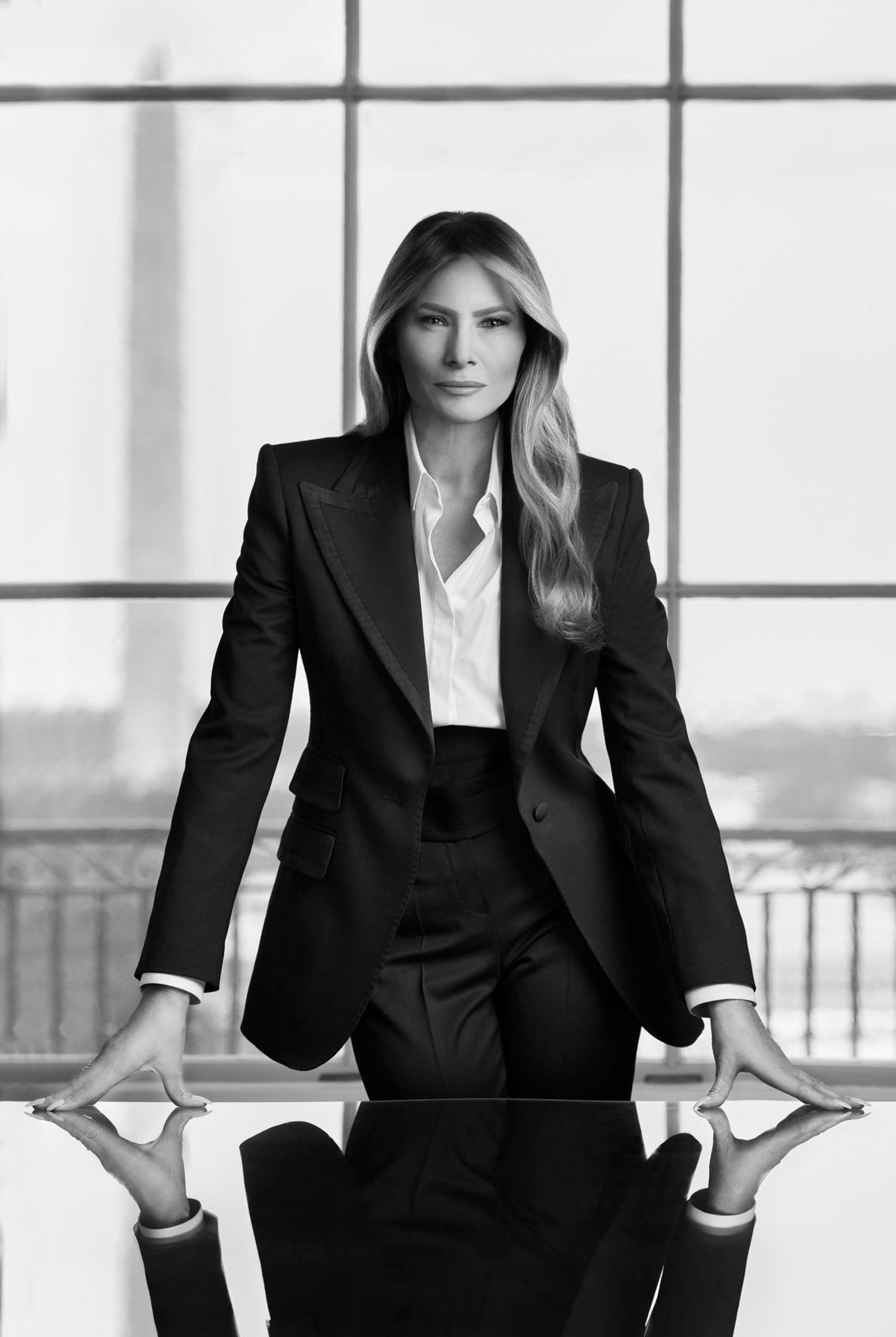 Melania Trump with her hands on the desk in a black and white suit with the Washington monument behind her