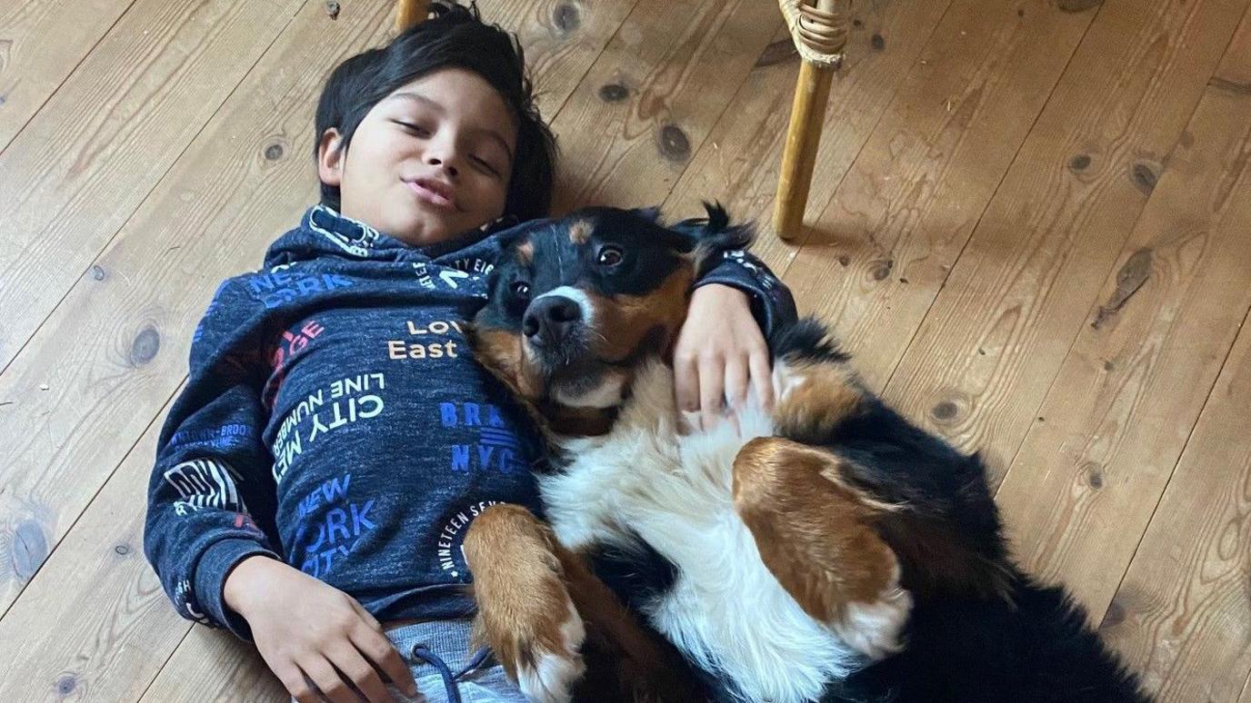 A young boy lying on the floor with his arm around Mali the dog