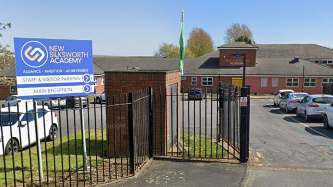 Black fencing in front of the school building which is made of brown brick and surrounded by a car park. There is a blue sign reading New Silksworth Academy. It points towards the Staff and Visitor Parking and the Main Reception.