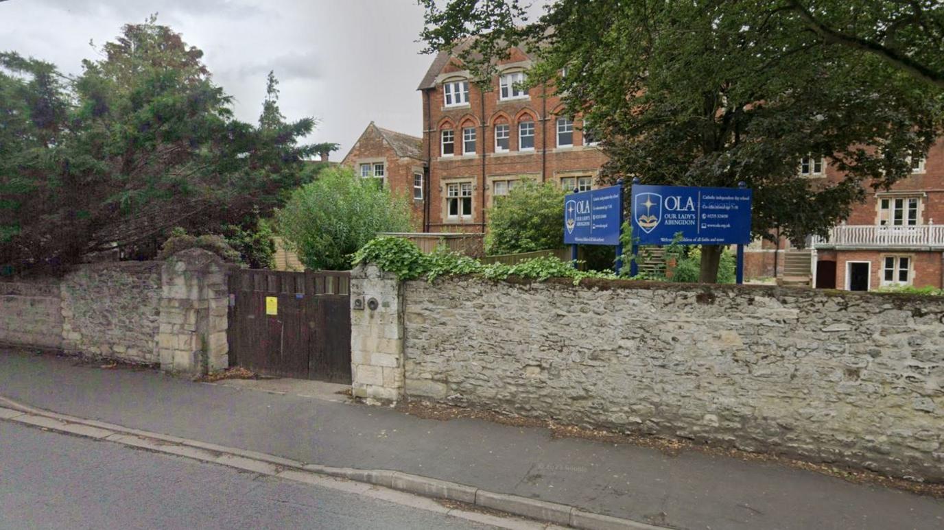 A Google Maps image of the front entrance to Our Lady's Abingdon. The school is a large red brick building partially hidden by a large tree. A wall has the school's sign on top which is blue. 