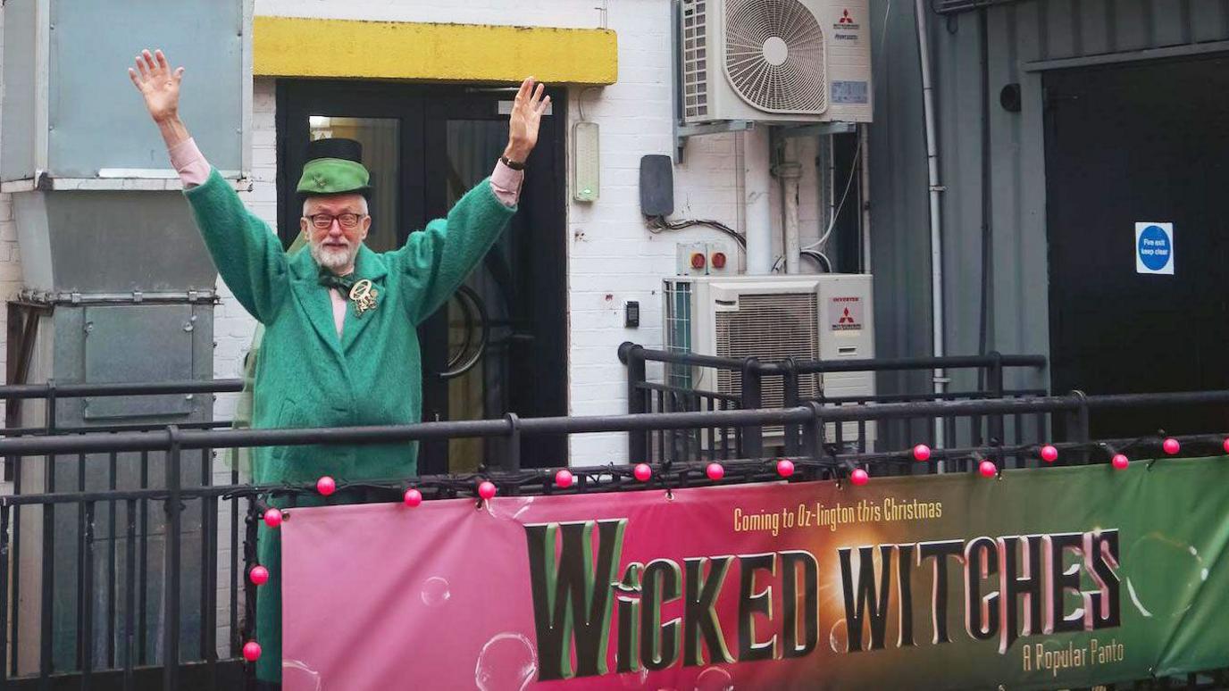 MP Jeremy Corbyn standing outside the Pleasance Theatre. He is wearing a green jacket and green hat and holding both his arms aloft. Behind him is a door and wall-mounted air conditioning units. There is a metal fence in front of him on which is displayed a banner advertising the Wicked Witches pantomime