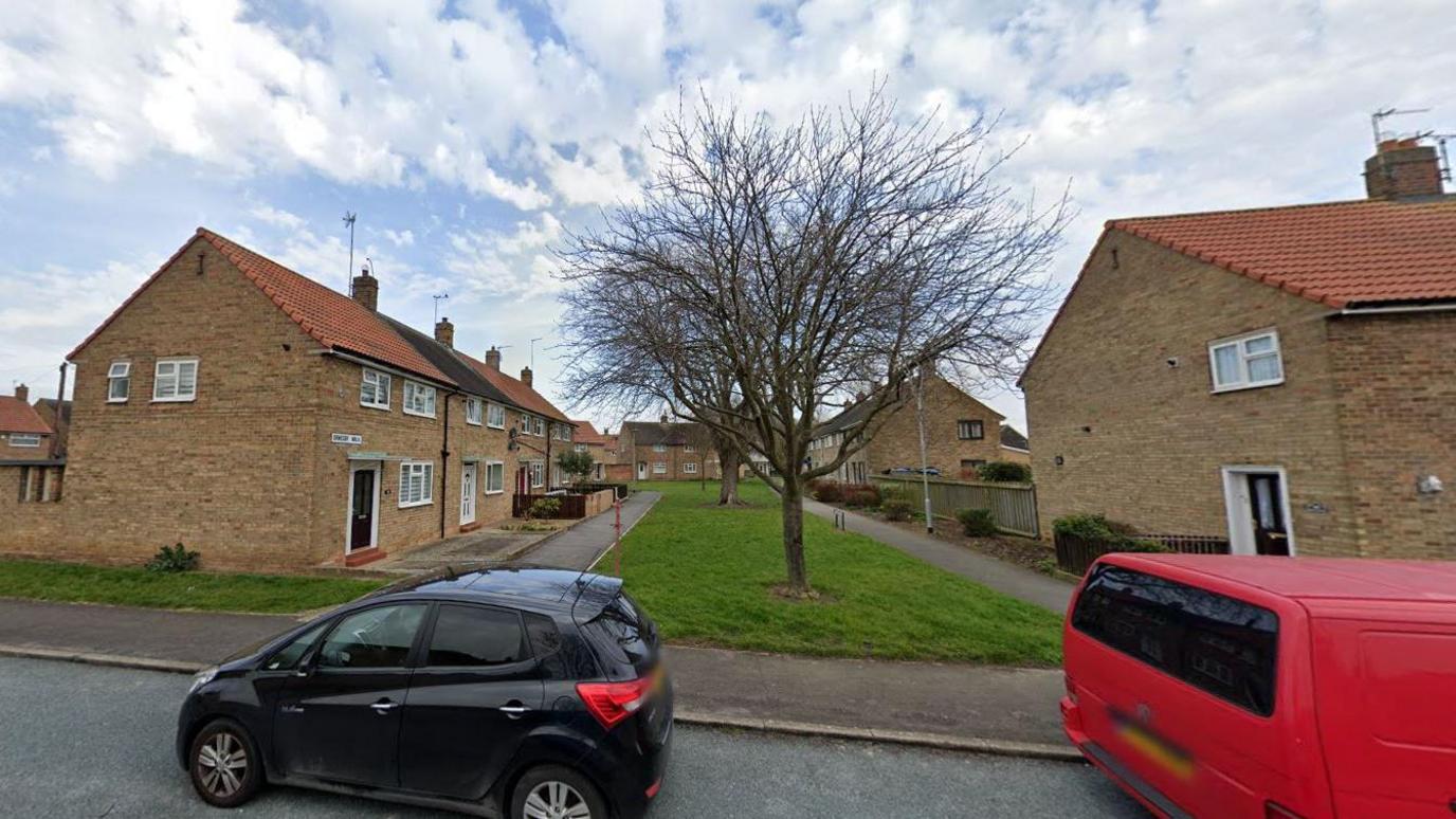 Google street view of the road facing Ormesby Walk which is a pathway with grass in the middle. There are several terraced houses along the path and two cars parked out front. 