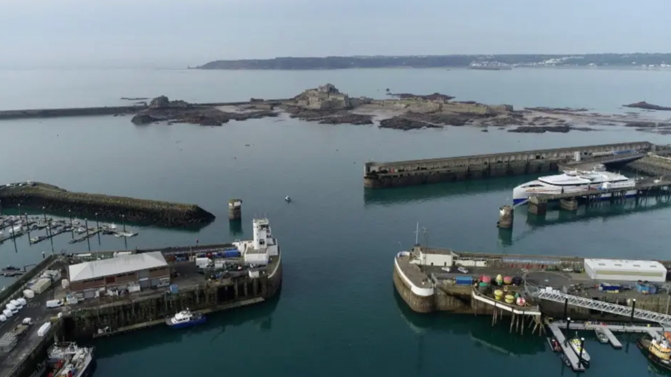 An aerial view of the Ports of Jersey - four separate walls sticking out in the water with boats surrounding, in the background you can see Elizabeth Castle and further afield is the coastline of Jersey.
