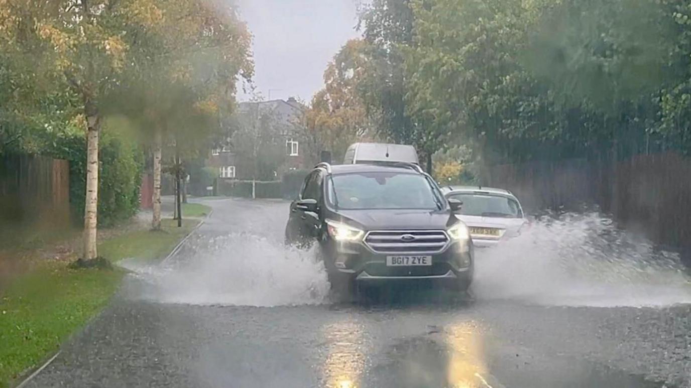 A car passes a parked vehicle in a flooded suburban street, with trees lining both sides