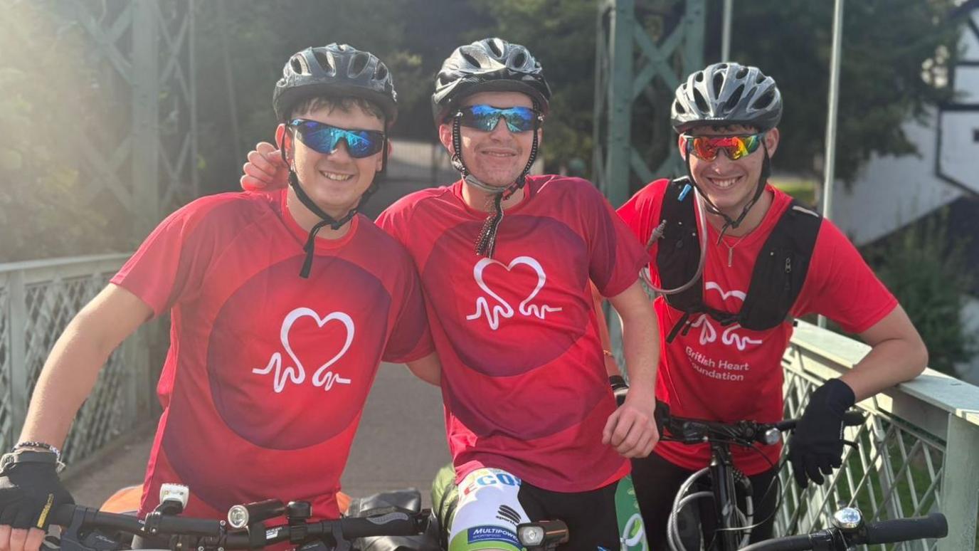 Dyfan Hughes, 17, James Thomas, 18, and Louis Dennis, 18, smile at the camera. They have their arms around each other and are wearing red T-shirts with the British Heart Foundation logo on it. They have sports sunglasses and bike helmets on and are on their bikes.