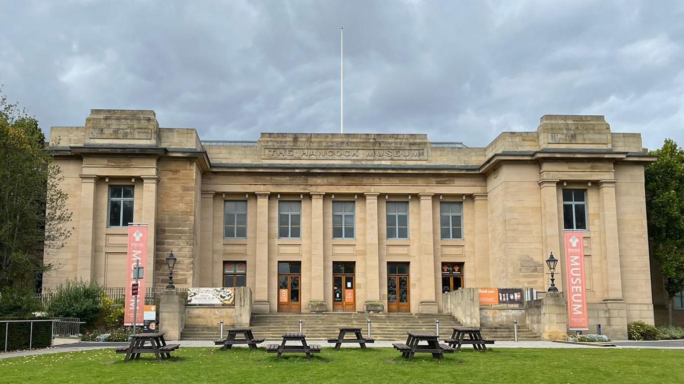 The Hancock Museum is a grand two-storey stone building. There are stone steps leading up to the wooden doors and there are picnic tables on the field out front. Orange banners either side of the building read: "Museum."
