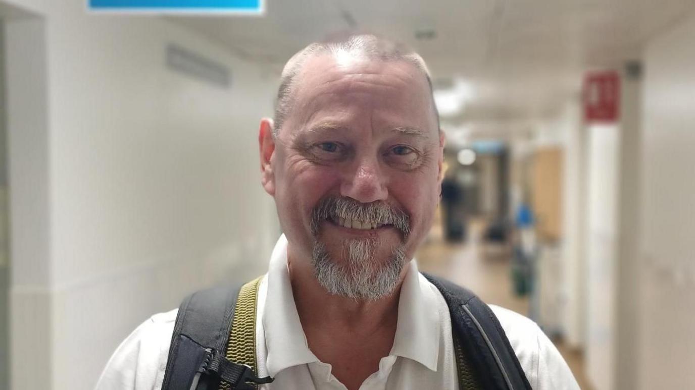 Ken Pottle is standing in a corridor in a hospital. He has short, thinning hair and a greying beard and moustache. He is wearing a white polo shirt and has a backpack over his shoulders
