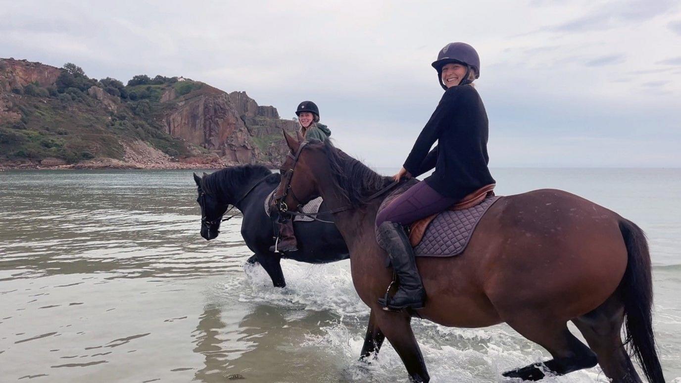 Two women on two horses in the sea. The first woman in the foreground is on a brown horse and  is wearing purple trousers, black boots, a black top and a purple helmet. The second woman is wearing a black helmet, green jacket and riding a black horse.