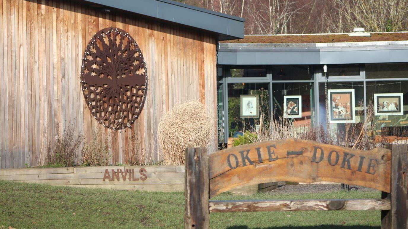 A close up of the Land of Oak & Iron heritage centre. A wooden bench is outside and iron artwork of an oak tree hangs on an outer wall. Paintings of dogs are displayed inside the centre, which can be seen through it's windows.