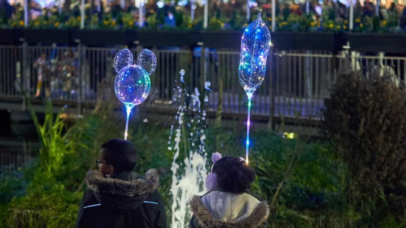 Two children holding light sticks, facing away from the camera at a town's light switch on. A boys is wearing a dark coat and a girl is wearing pink earmuffs and a coat with a fluffy hood. They are standing around trees and bushes. 