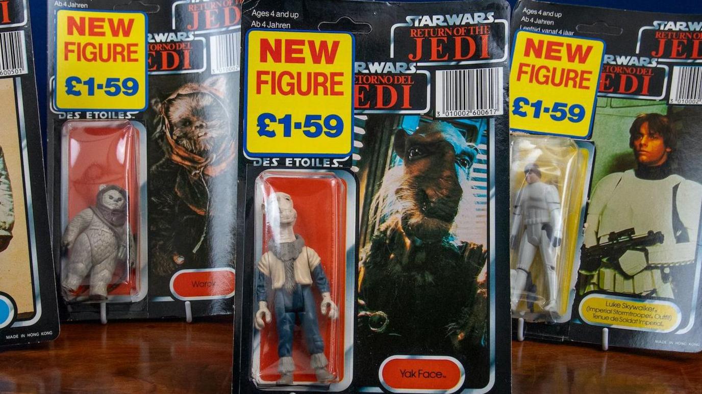 A mixture of five Star Wars figurines, all standing on a wooden table. all with £1.59 price tags on them. They are all in their original boxes.
