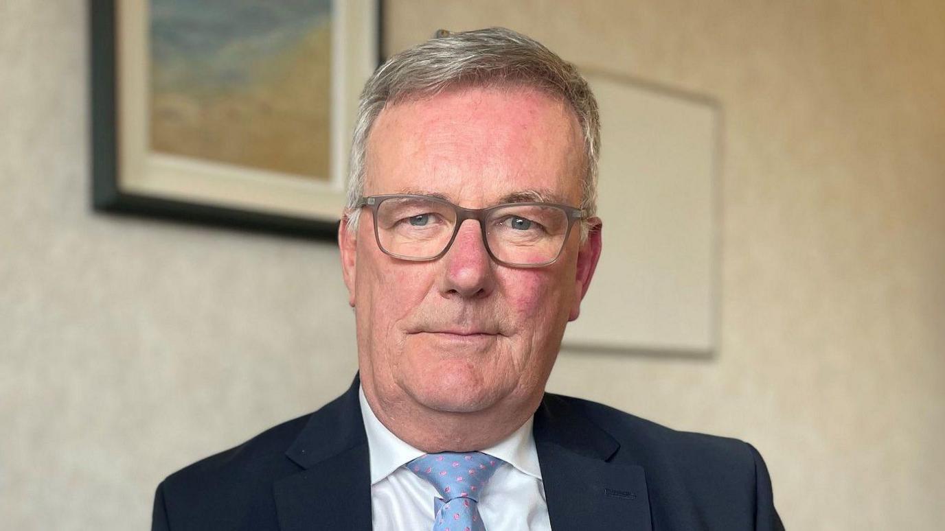 Mike Nesbitt wears a black suit, white shirt and blue tie. He is wearing glasses and has grey hair. He is looking at the camera. There are photos on the wall behind him. 