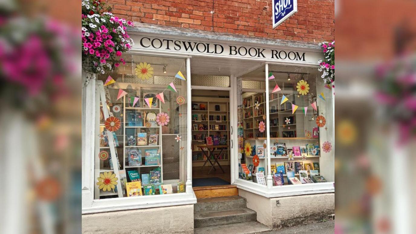 The outside of Cotswold Book Room on a warm sunny day. The door is open and there are lots of books on display, flower stickers and colourful bunting in the windows. There are hanging baskets on either side of the sign above the door, with white and pink cascading flowers.